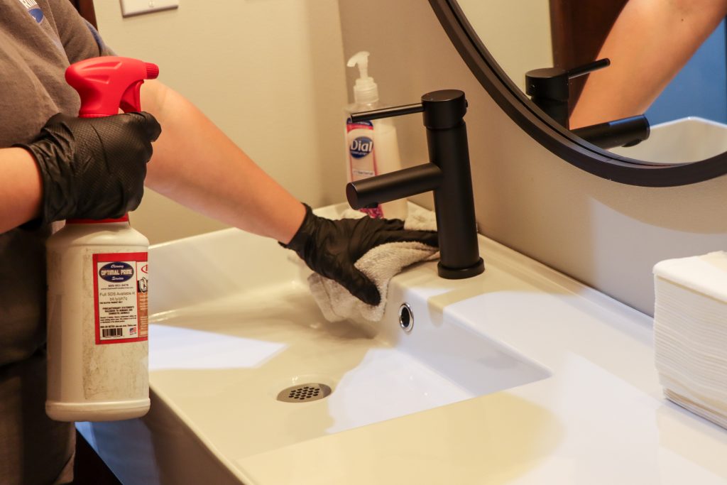 Person with black gloves cleaning a bathroom sink using a white cloth and cleaning spray, with a black faucet and a mirror in the background.