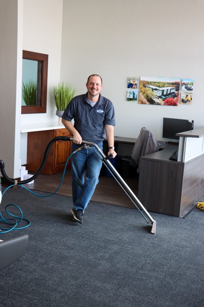 A man cleaning a carpet in an office with a carpet cleaning machine, surrounded by office furniture and decor.