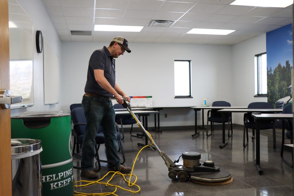 Person operating a floor cleaning machine in an office with tables, chairs, and a mural on the wall.