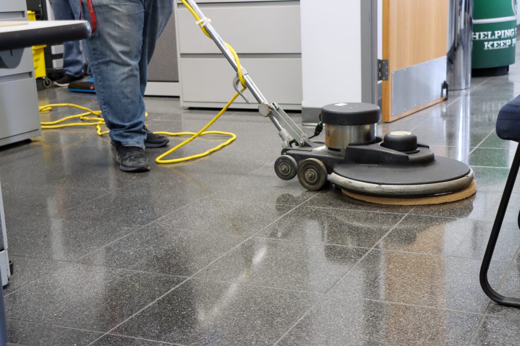 Person using a floor polishing machine on a dark tiled floor in an office, with a yellow cord and office furniture in the background.