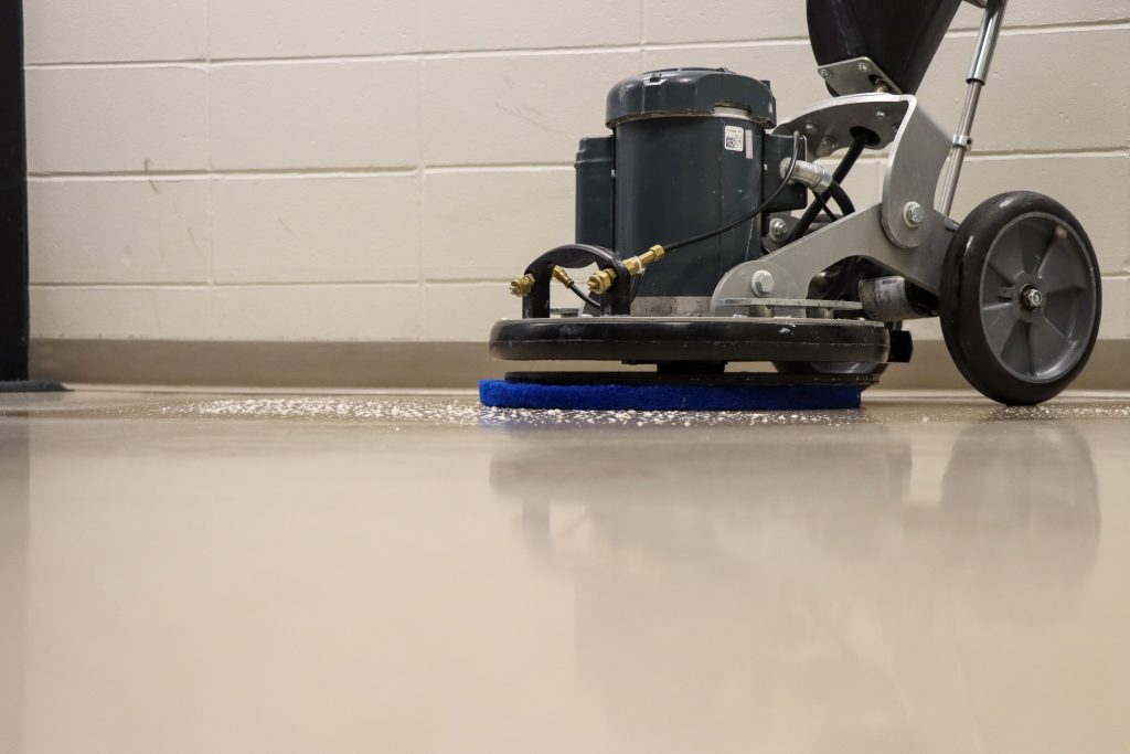 Close-up of a floor polishing machine on a concrete floor with a tiled wall in the background.